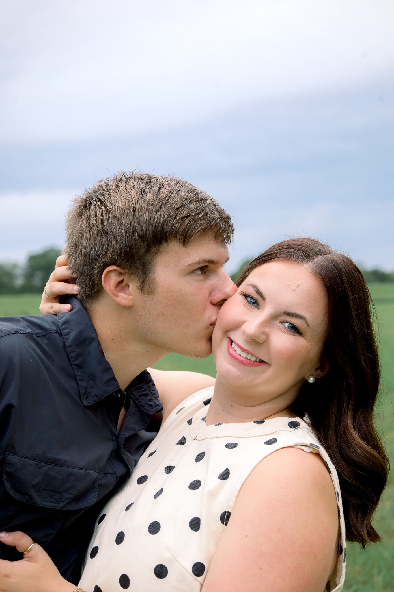 A young man kisses a smiling woman on the cheek outdoors in a green field under a cloudy sky.