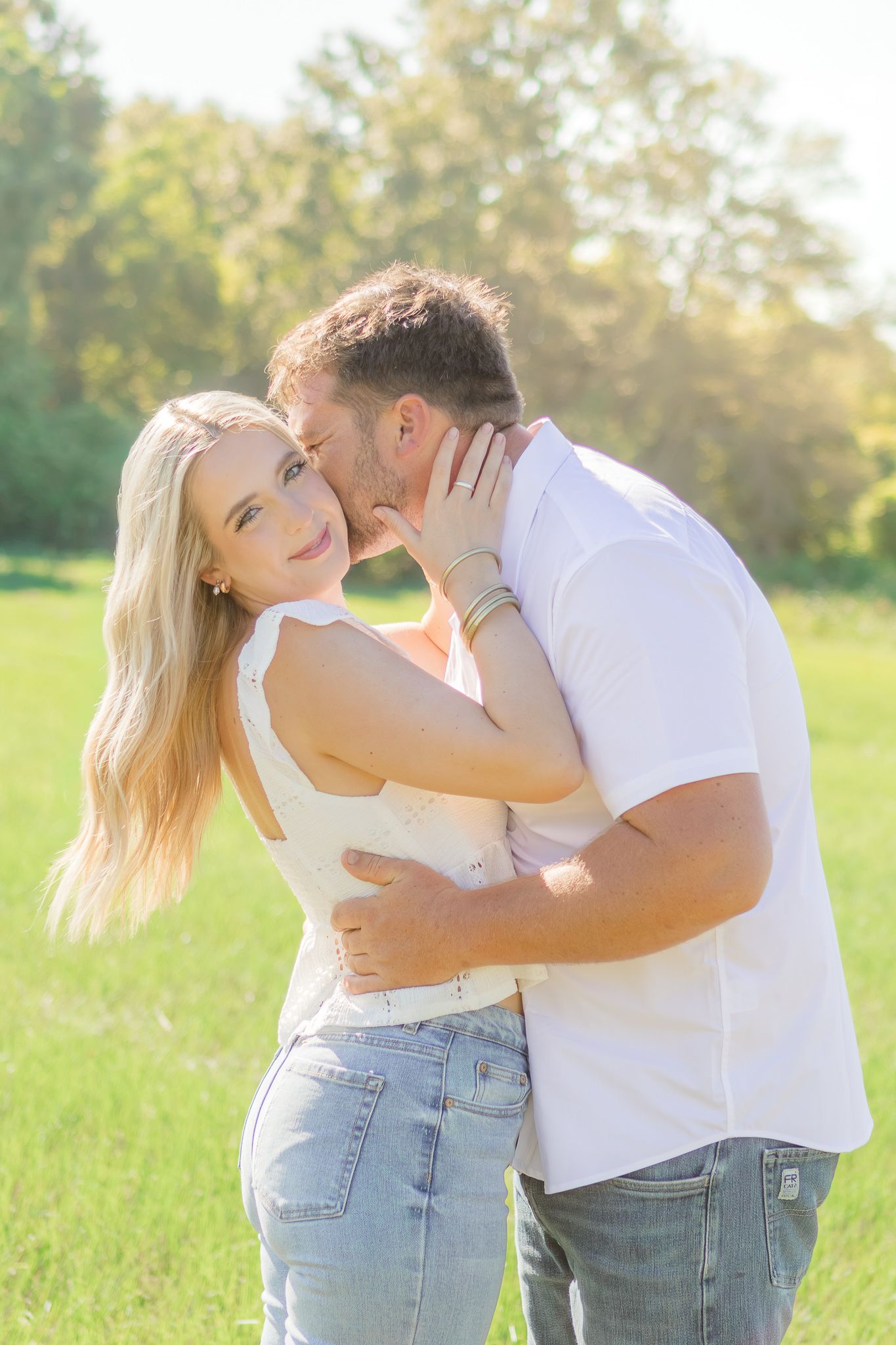 A couple shares a tender moment outdoors, man kissing womans cheek in a sunny green field.