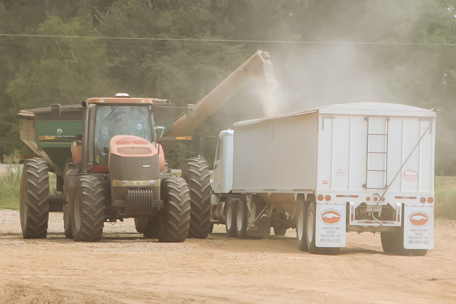 Red tractor using grain auger to transfer harvested crops into a white trailer on a farm.