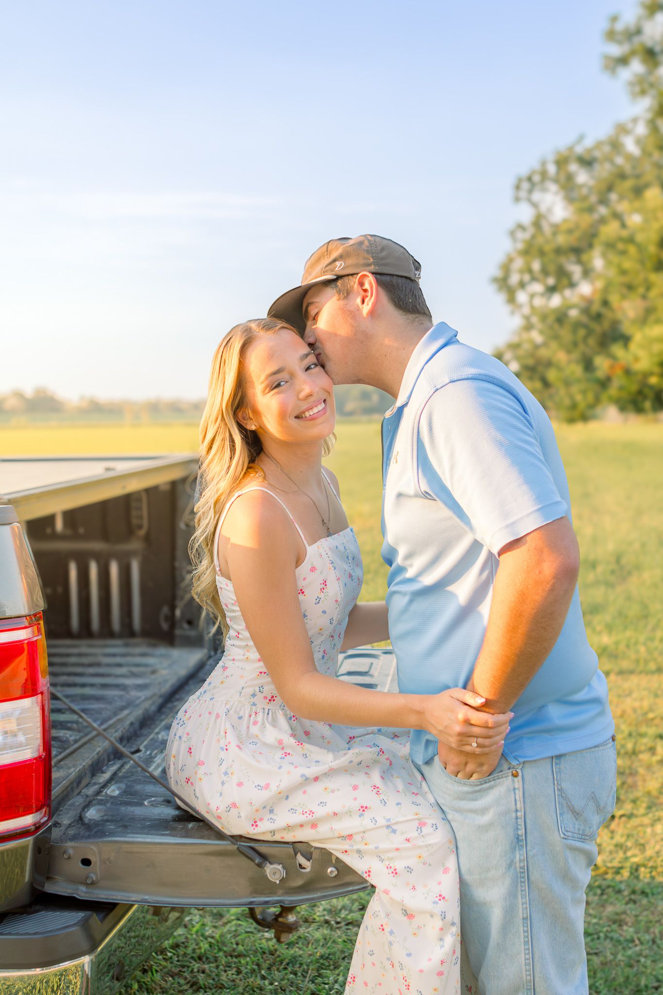 Couple sharing a romantic moment on a red truck tailgate in a sunny field, woman in floral dress smiling.