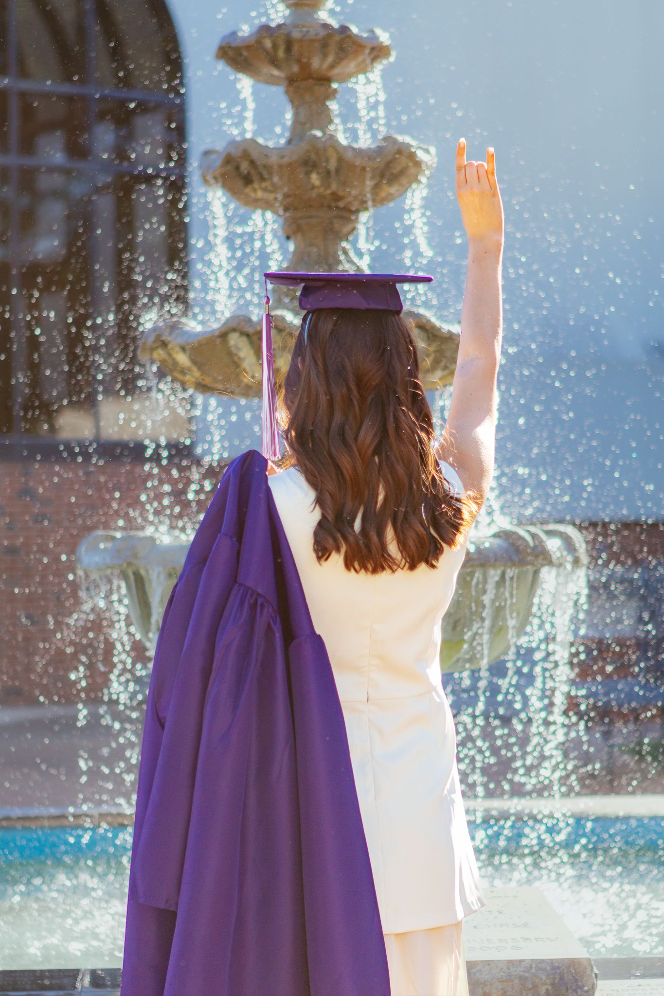 Graduate in white dress and purple cap raising hand toward an ornate stone fountain on campus.