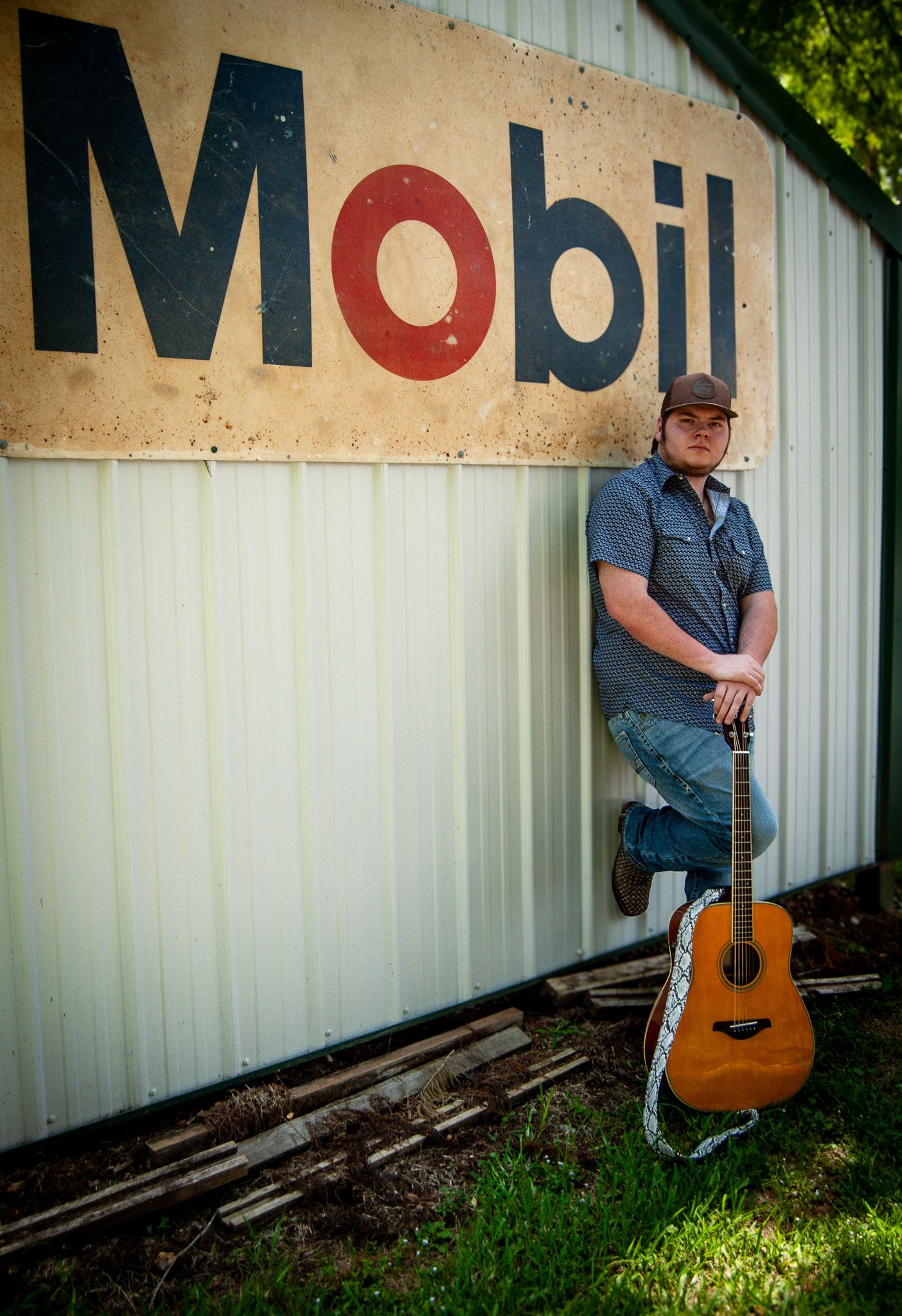 Musician with acoustic guitar posing against industrial wall with Mobil sign.