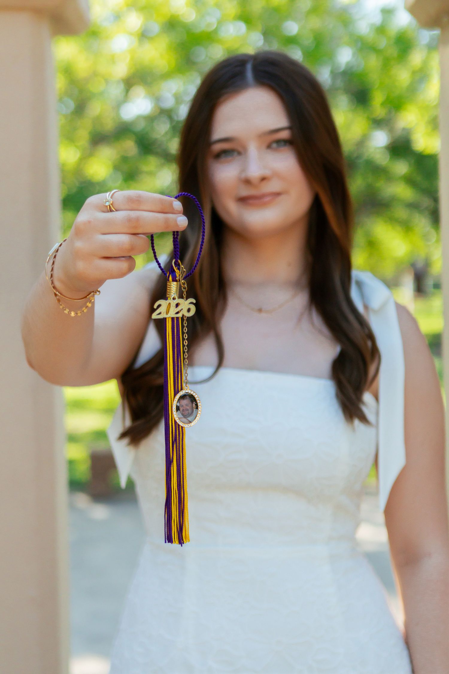 Senior girl in white dress holding 2026 graduation tassel charm toward camera in outdoor portrait.