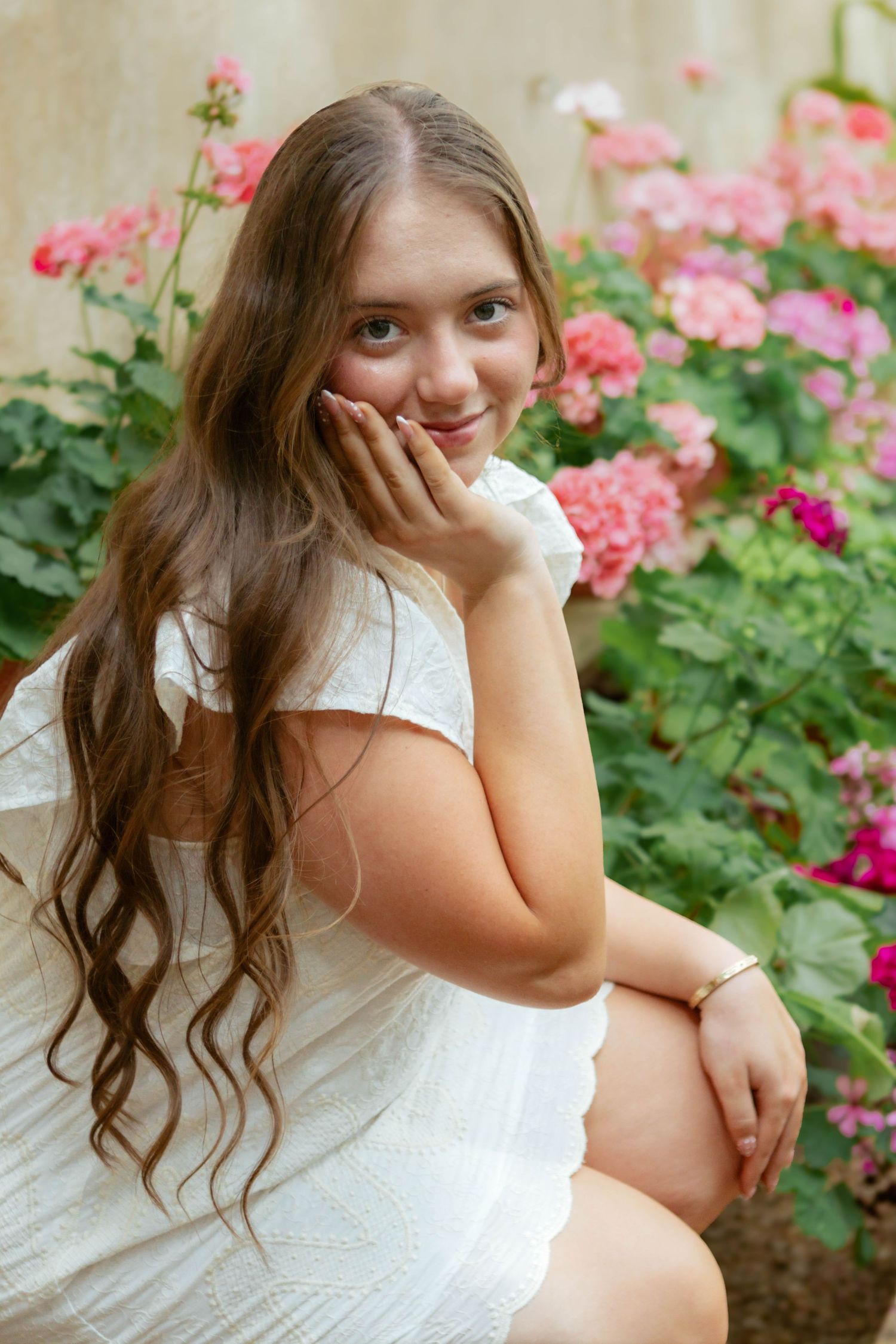 Teenage girl with long wavy hair in white dress sitting among pink flowering bushes smiling at camera.