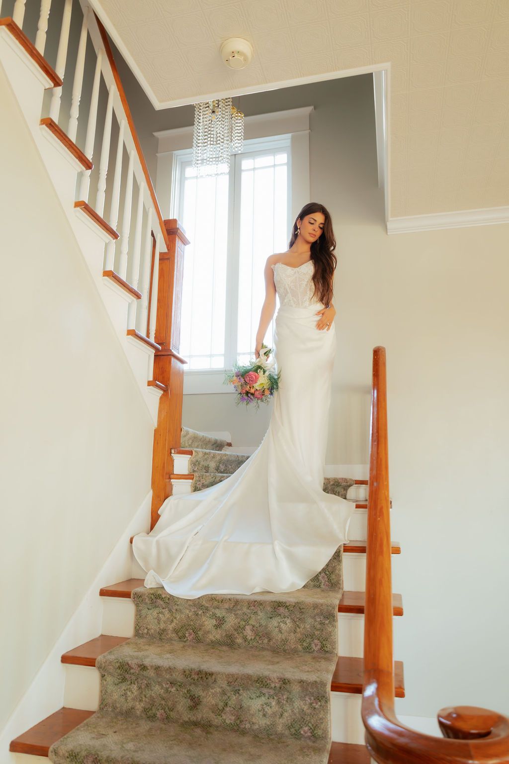 Brunette bride in a flowing white satin gown with train descending a classic wooden staircase holding bouquet.