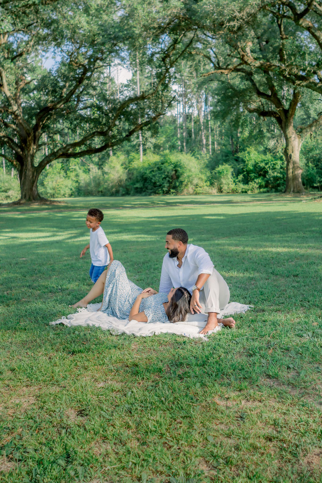 Family relaxing on a blanket in a lush green park, with a child running nearby under large oak trees.