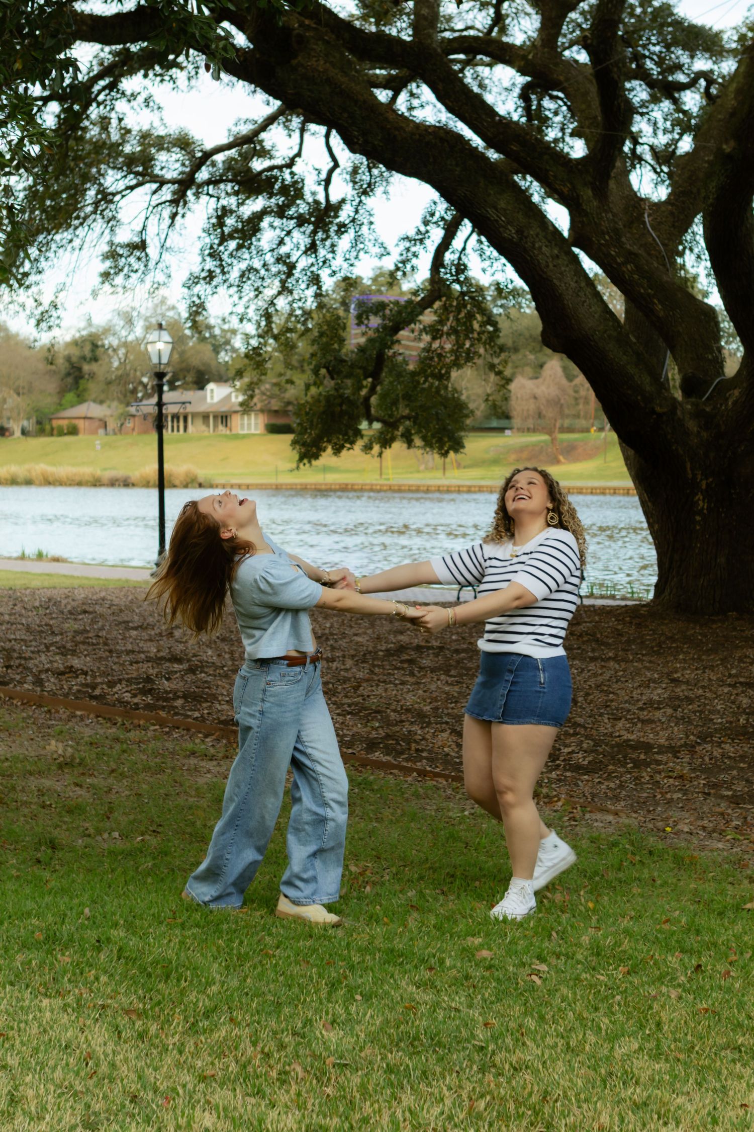 Two friends laugh and play together under a large oak tree by a serene lake on a sunny afternoon.