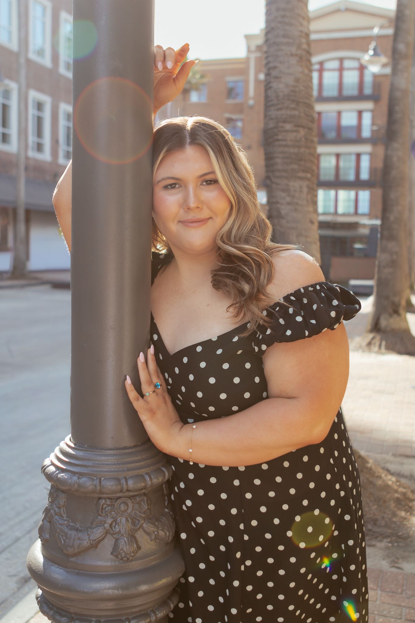 Young woman in black polka dot off-shoulder dress posing against a decorative street lamp in an urban setting.