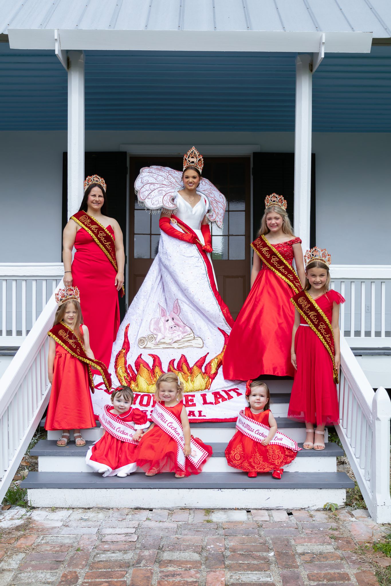 Beauty pageant queens in red gowns and crowns pose on porch steps at Cochon de Lait Festival.