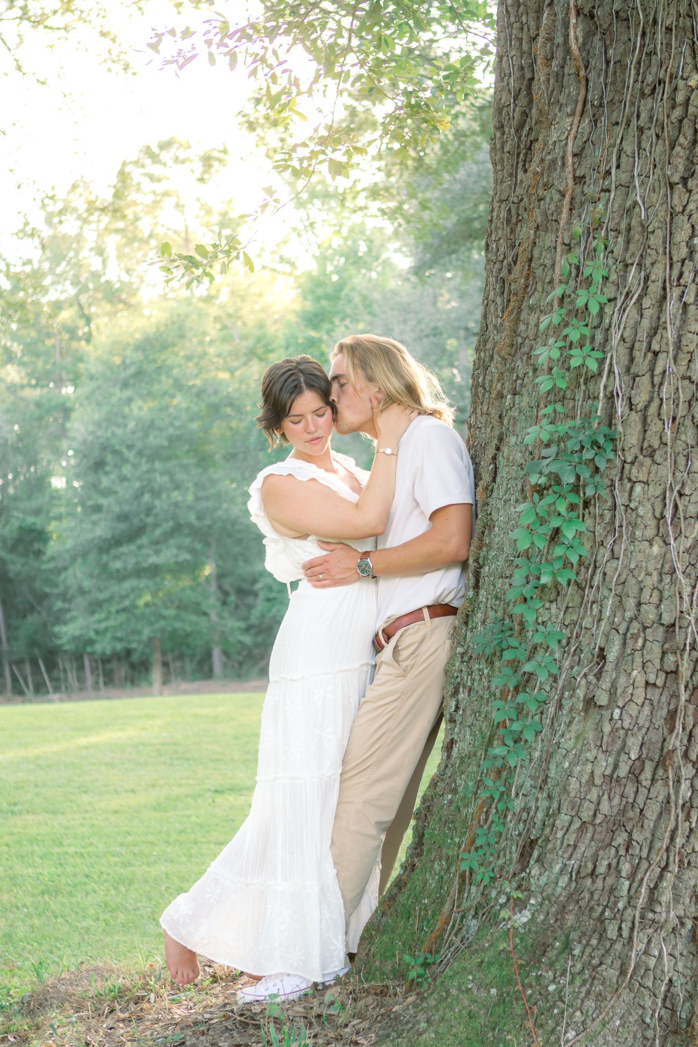 Couple sharing a romantic kiss by a large tree in a lush green park, woman in white dress.