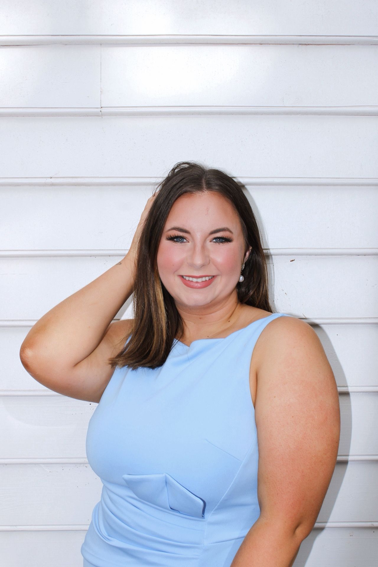 Smiling brunette woman in a light blue sleeveless dress posing against a white paneled wall.