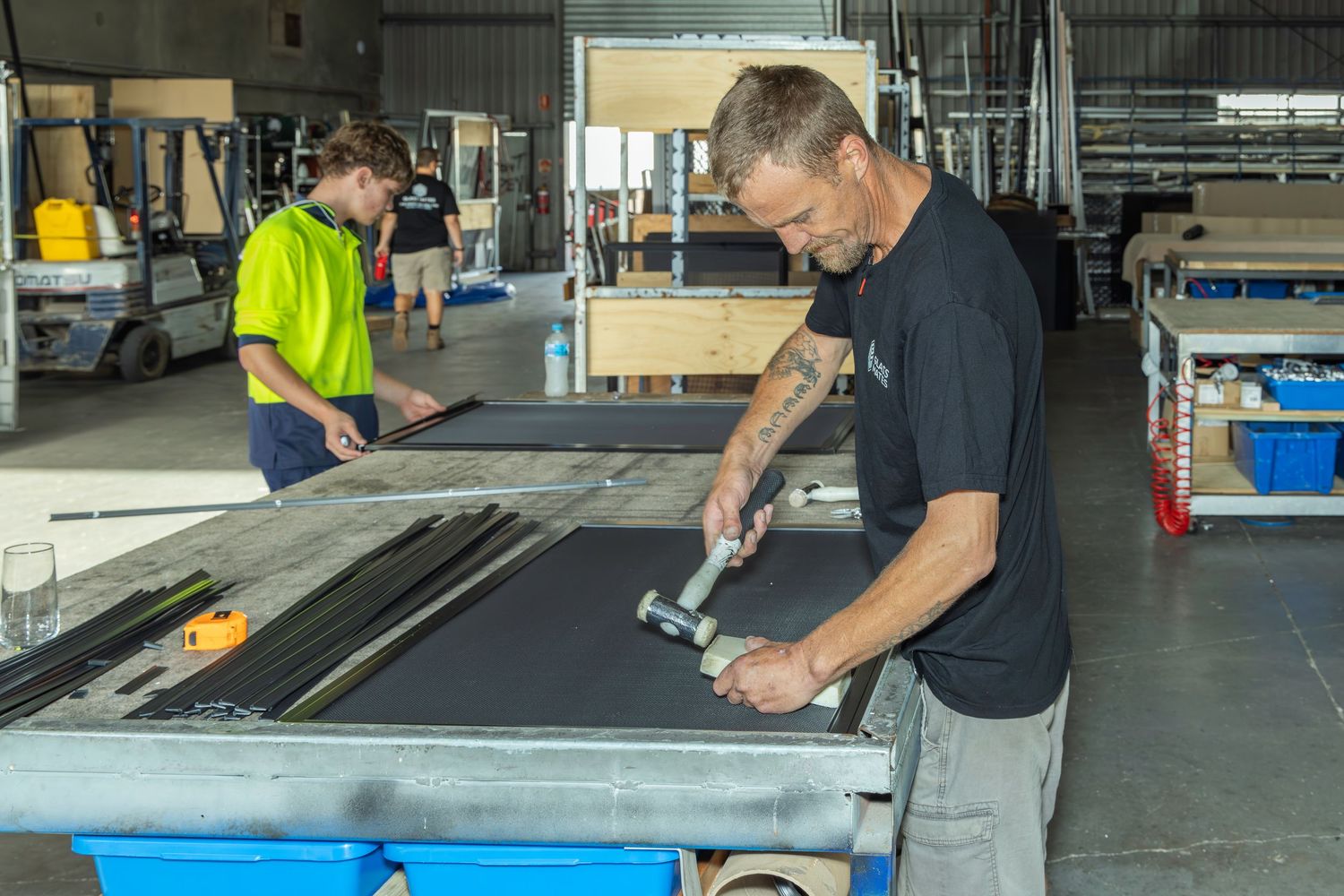 Two workers assembling dark-framed panels in an industrial workshop using hand tools.
