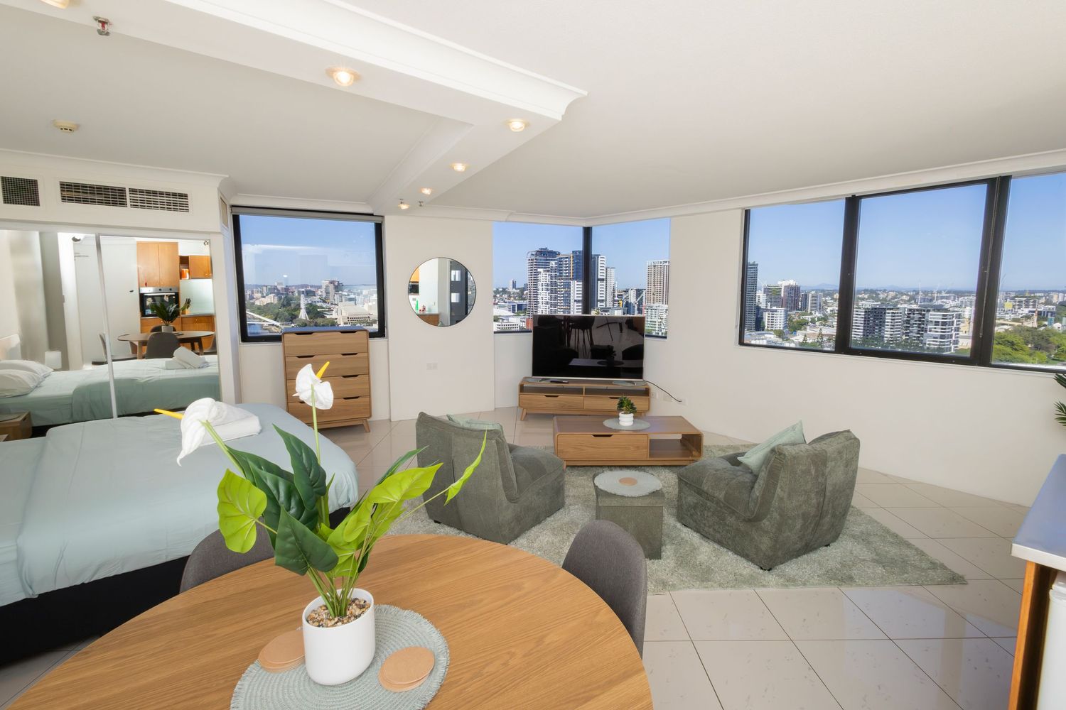 Modern high-rise apartment living room with city skyline views, featuring green sofas, wooden furniture, and a flat-screen TV.