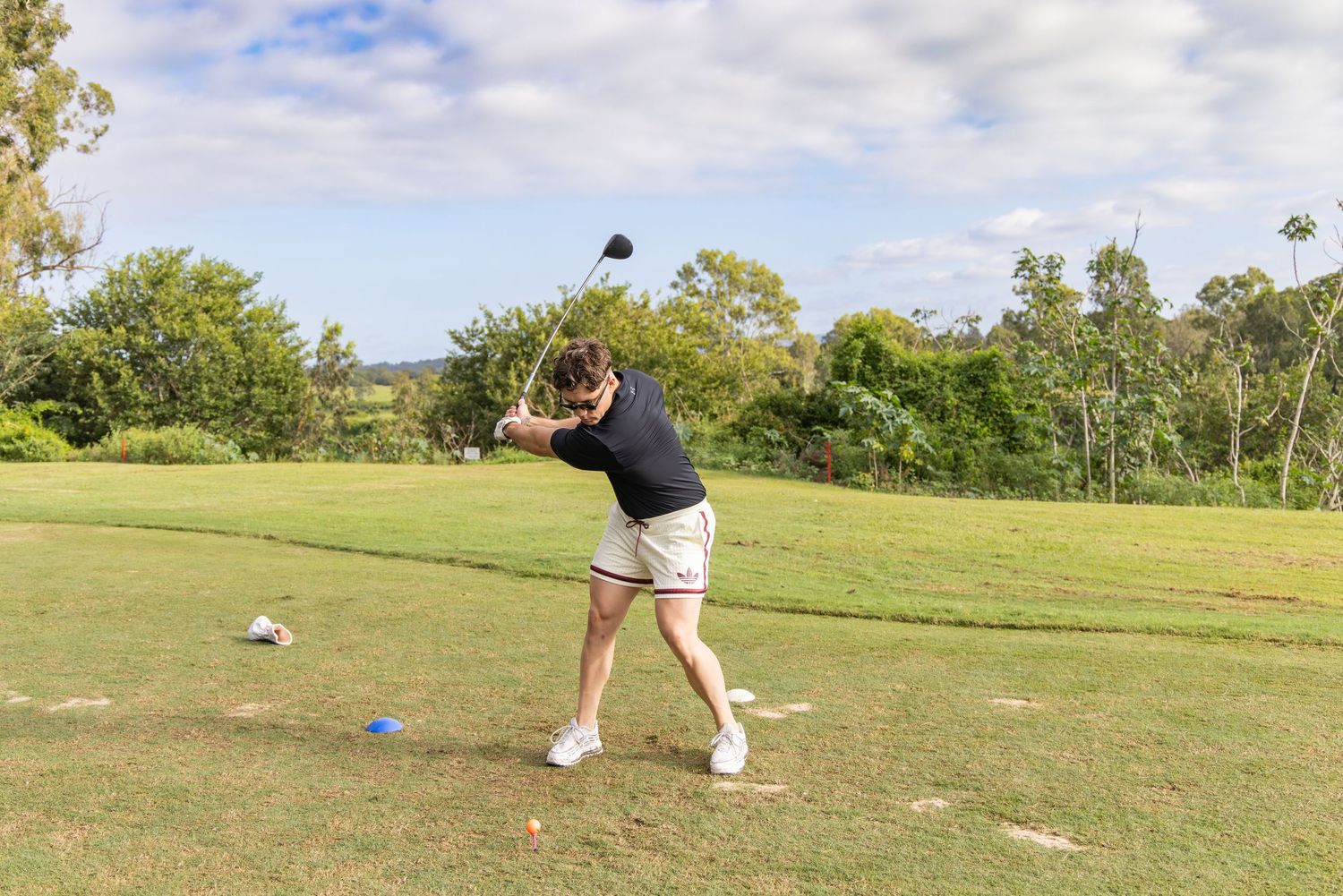 Golfer mid-swing on a lush green golf course with tropical trees and blue sky in the background.