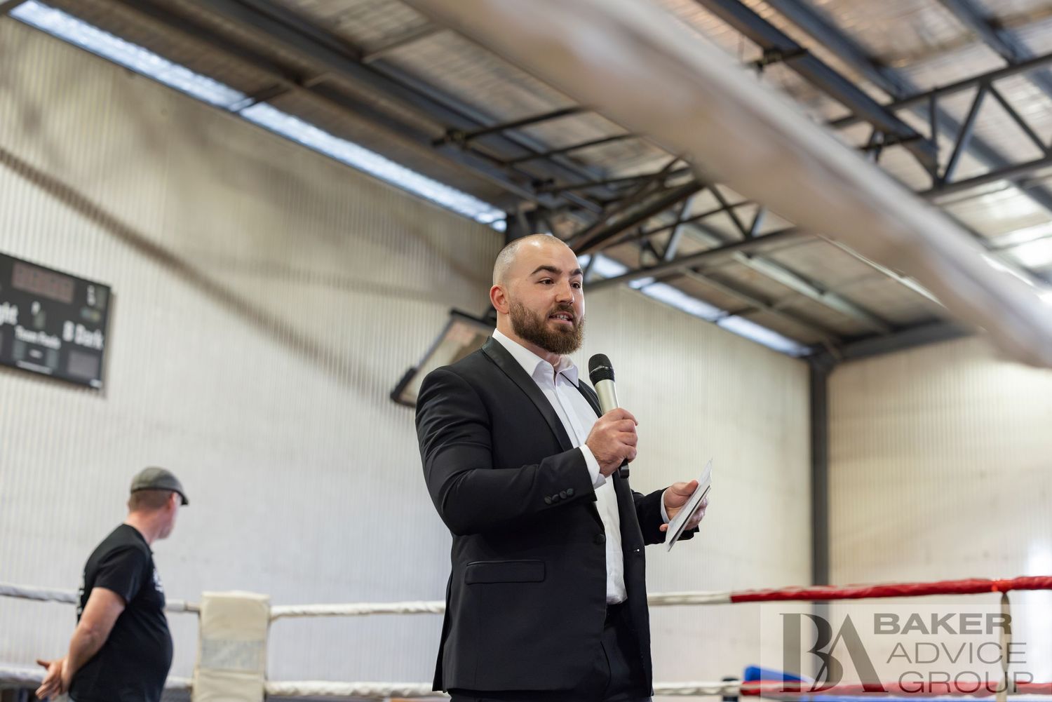 Man in black suit speaking into microphone at boxing ring event with Baker Advice Group branding visible.