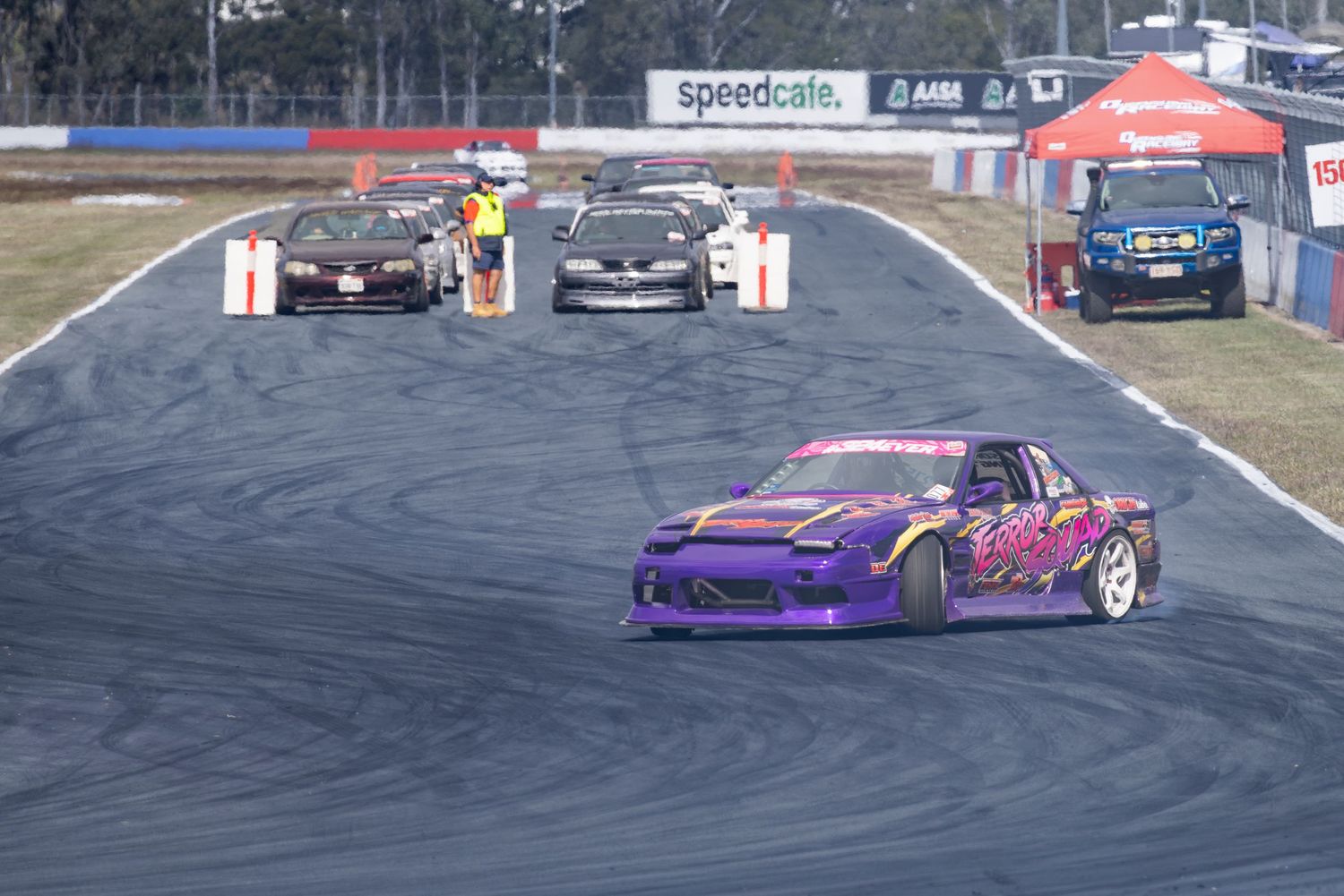 Purple drift car leads a pack of race cars around a track corner at a Speedcafe motorsport event.