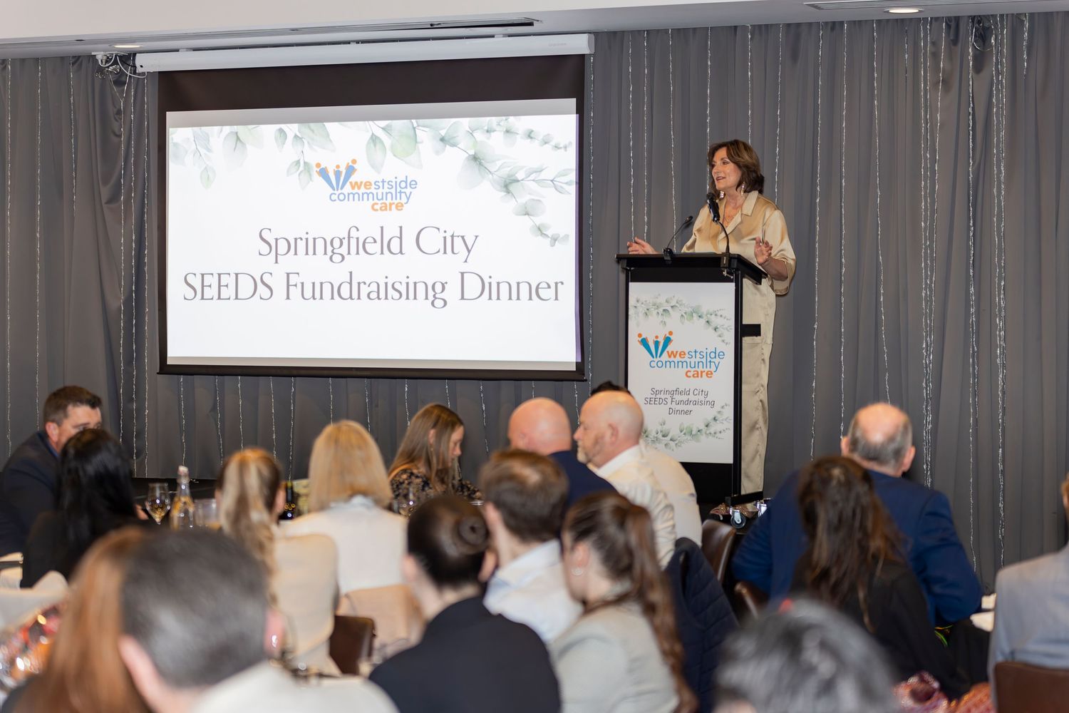 Woman speaks at podium during Springfield City SEEDS Fundraising Dinner event with attendees seated at tables.