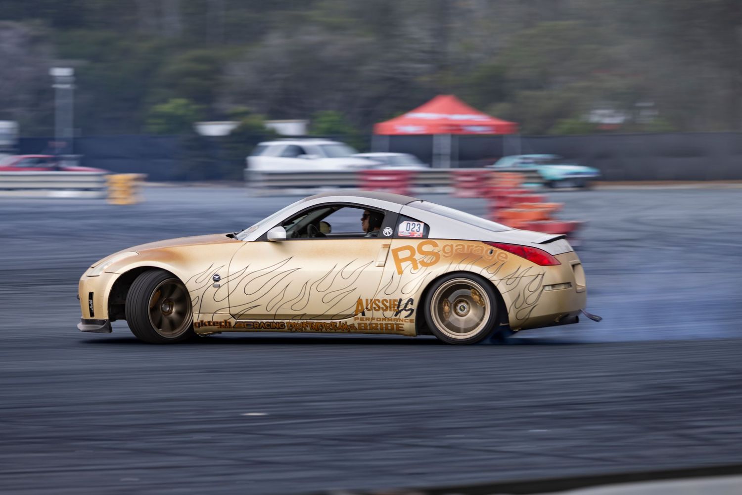 Gold Nissan 350Z drifting on a race track, leaving tire smoke trails during a motorsport competition.