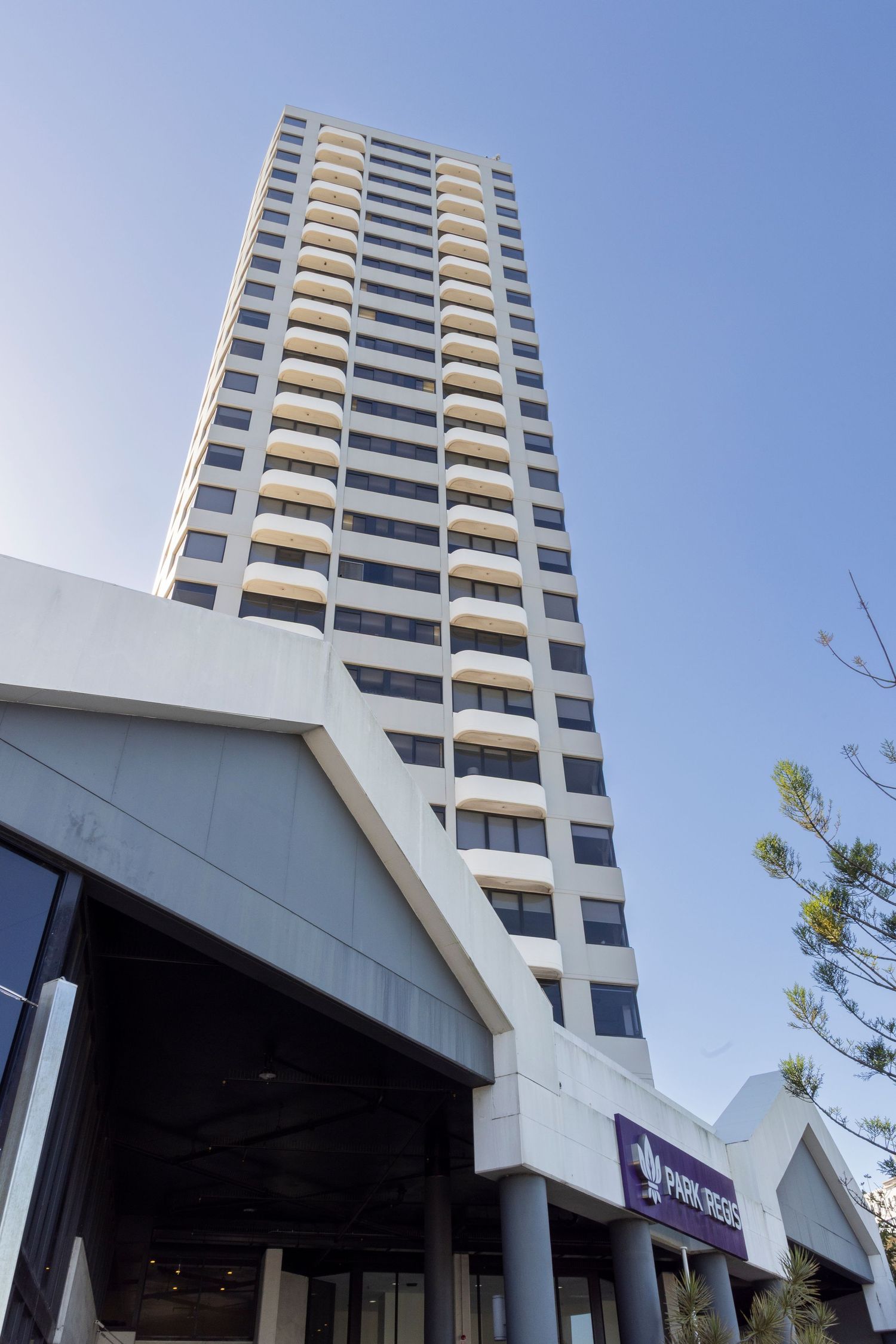 Tall modern high-rise hotel building with Park Regis sign at entrance against clear blue sky.