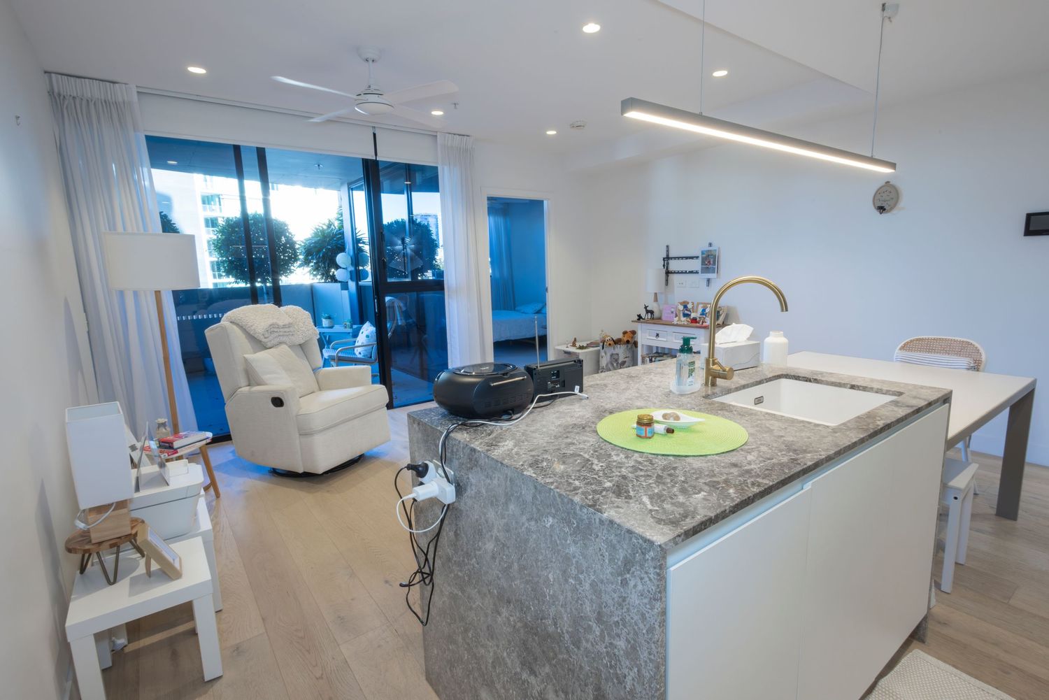 Modern open-plan kitchen with marble island, white interiors, and floor-to-ceiling windows leading to a bright living area.