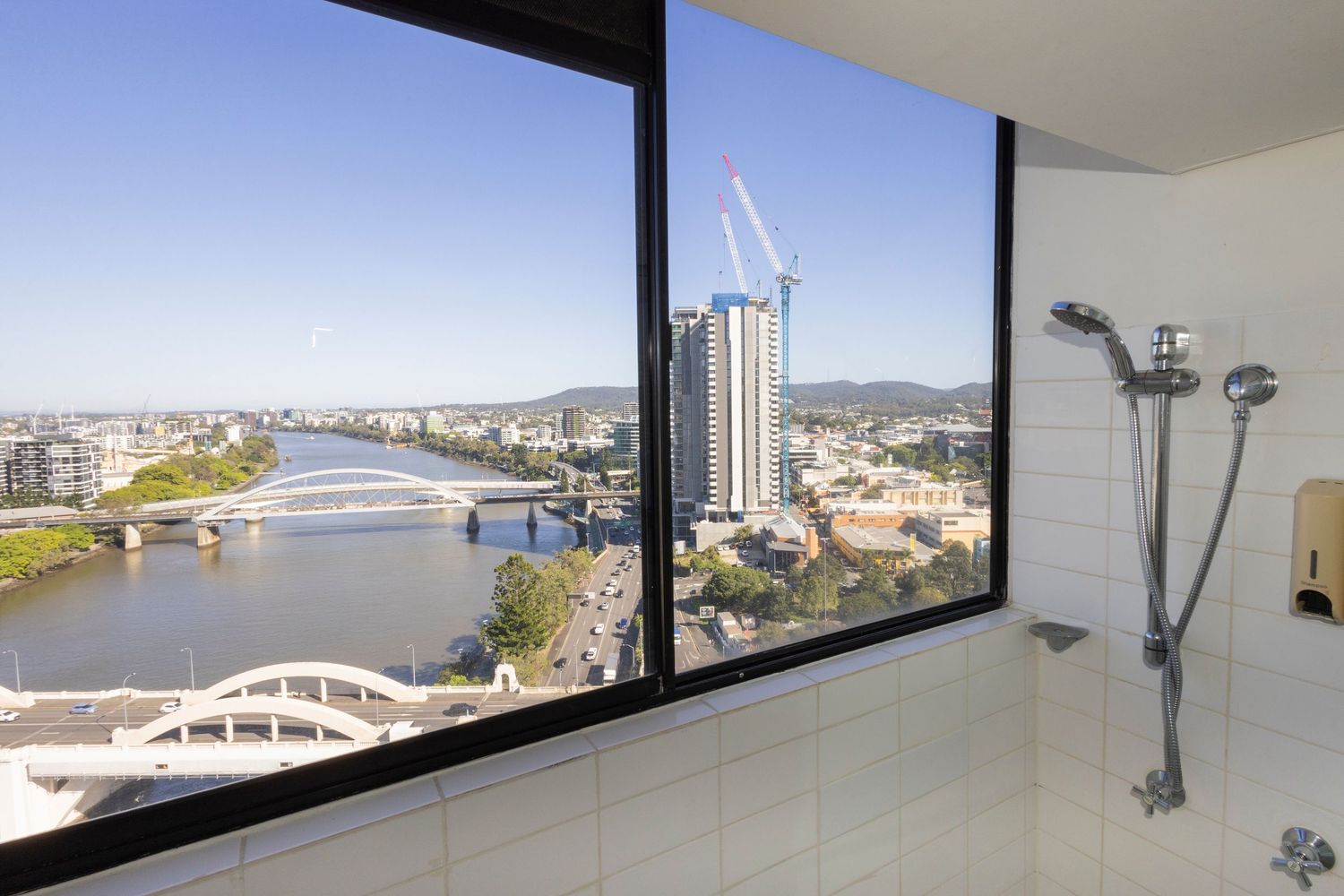 Luxury bathroom with large window overlooking Brisbane River, bridge, and city skyline with a walk-in shower.