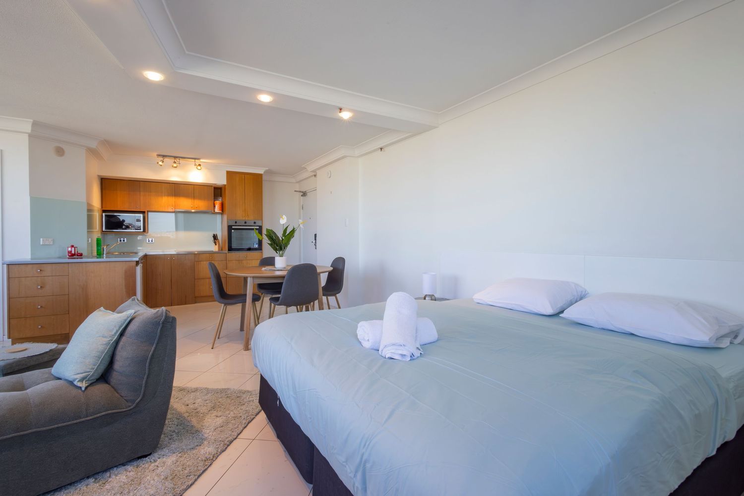 Modern studio apartment with a king bed, gray armchair, and open-plan kitchen with wooden cabinets and dining area.