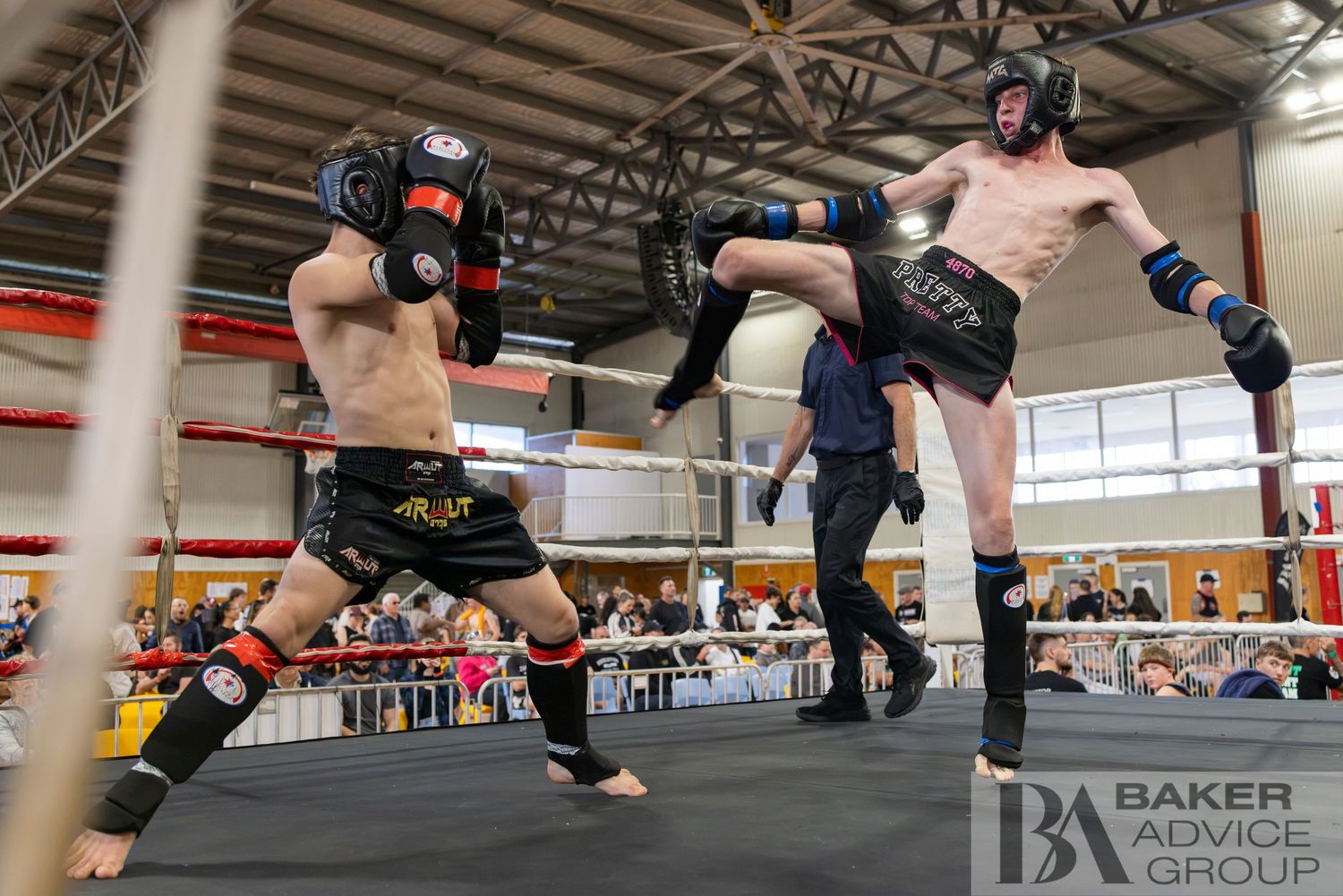 Two kickboxers sparring in a ring, one executing a high kick while wearing headgear and gloves at an indoor competition.
