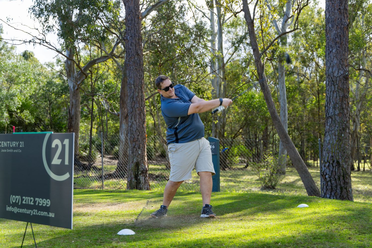 Man swinging a golf club on a lush green course near trees, with a Century 21 real estate sign nearby.