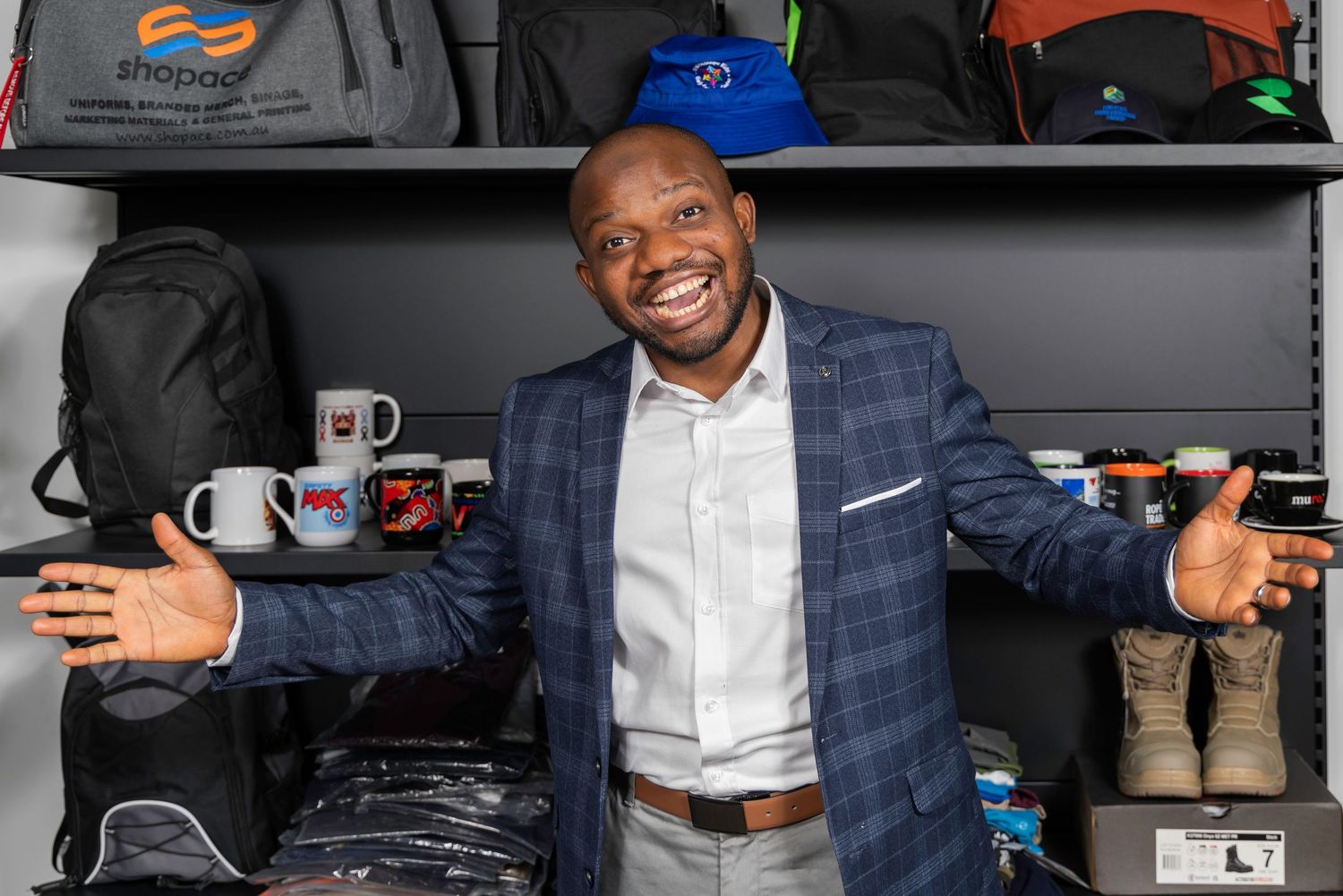 Smiling businessman in blue blazer poses among branded merchandise, bags, mugs, and products on display shelves.