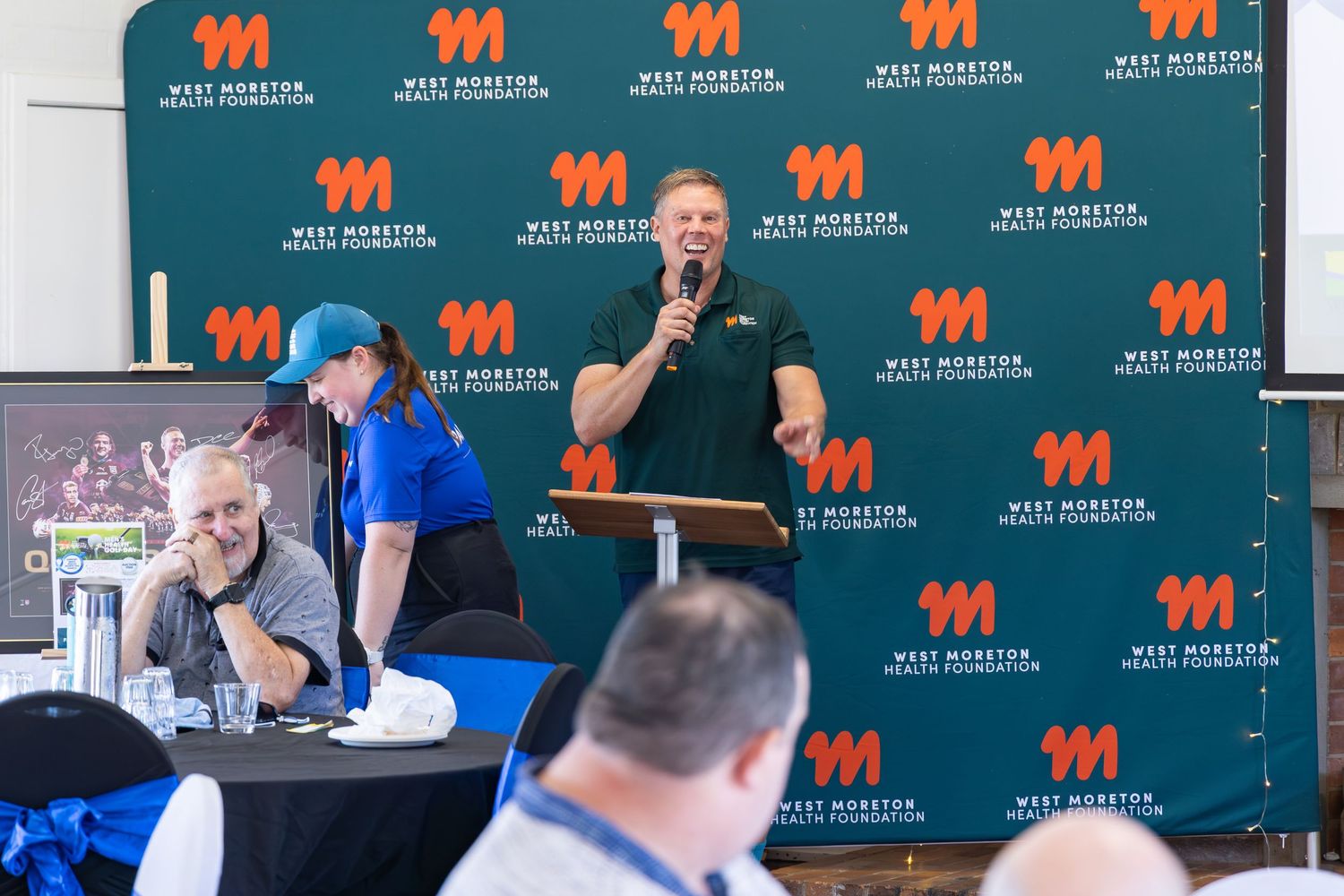 Man speaks into microphone at West Morton Educational Foundation event, with attendees seated at tables in foreground.