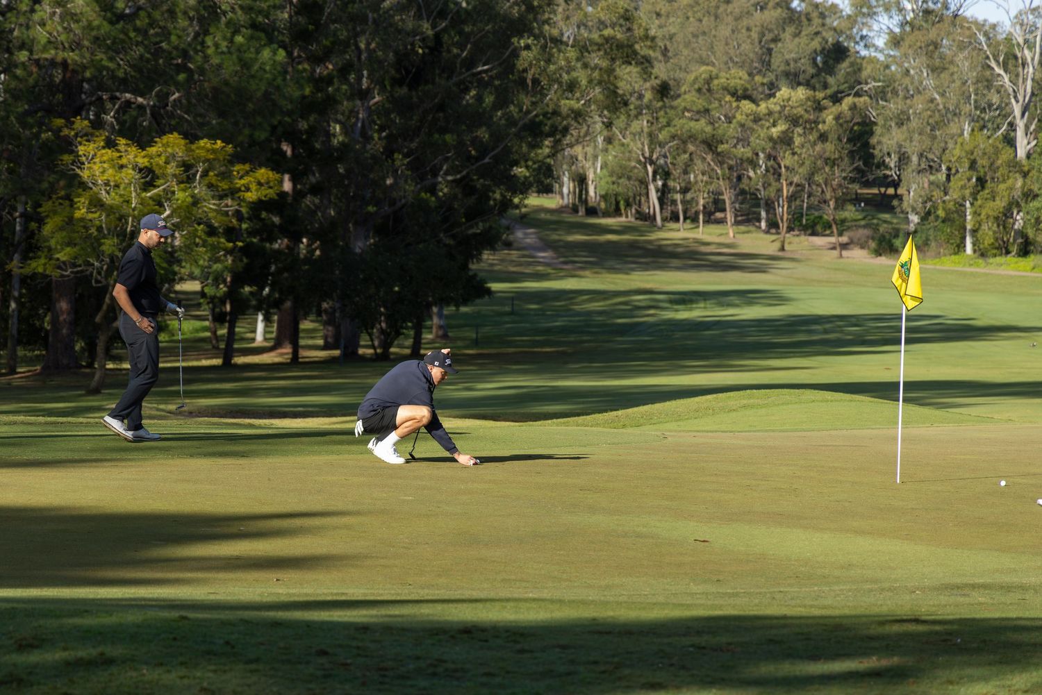 Two golfers on a lush green golf course, one crouching to read the putt near a yellow flag.