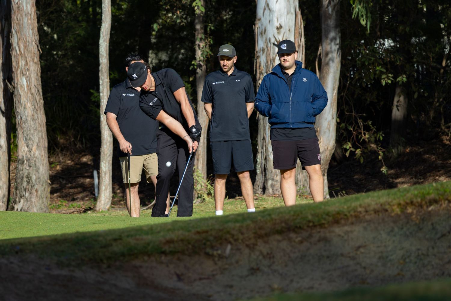 Four golfers in dark attire discuss strategy near a sand bunker surrounded by tall pine trees on a sunny golf course.