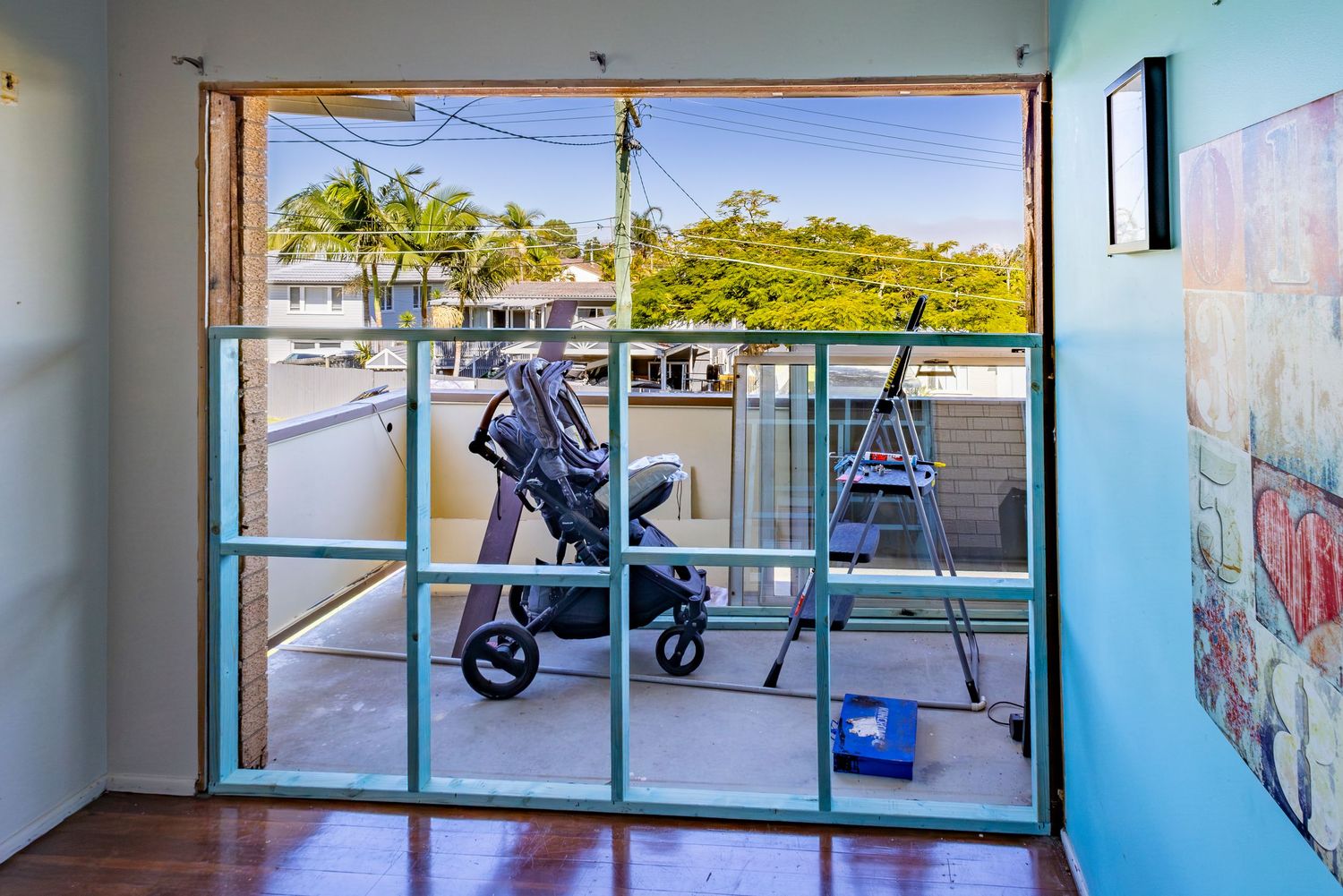 Glass sliding doors open to a balcony with a stroller and ladder, overlooking a tropical street with palm trees.