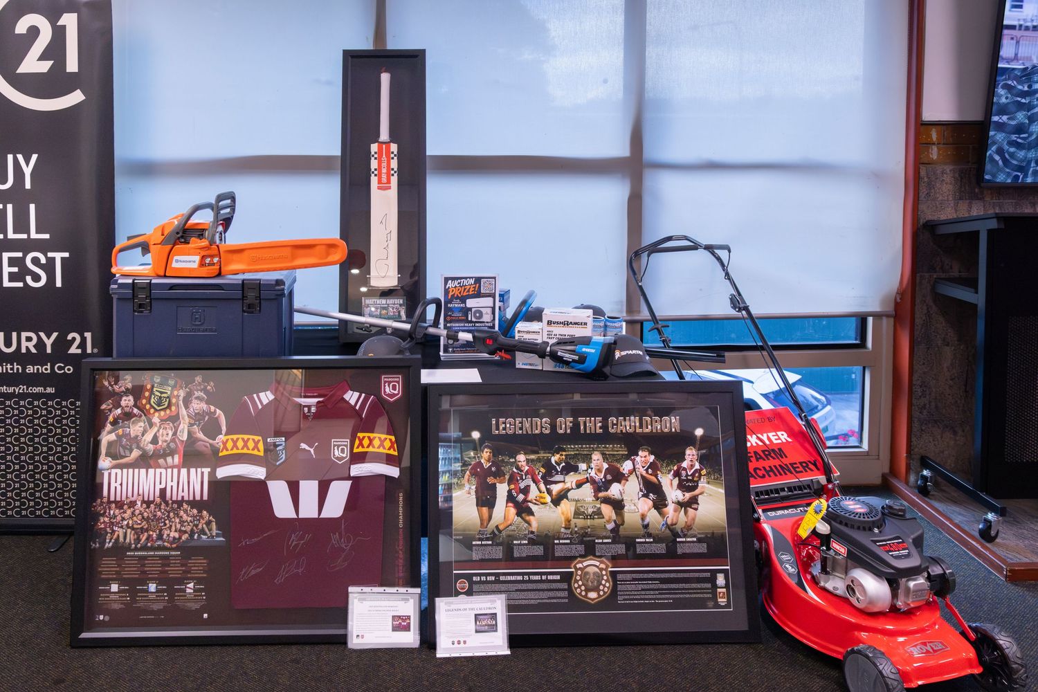 Display of auction items including a chainsaw, cricket bat, framed sports memorabilia, and red lawn mower at a Century 21 charity event.
