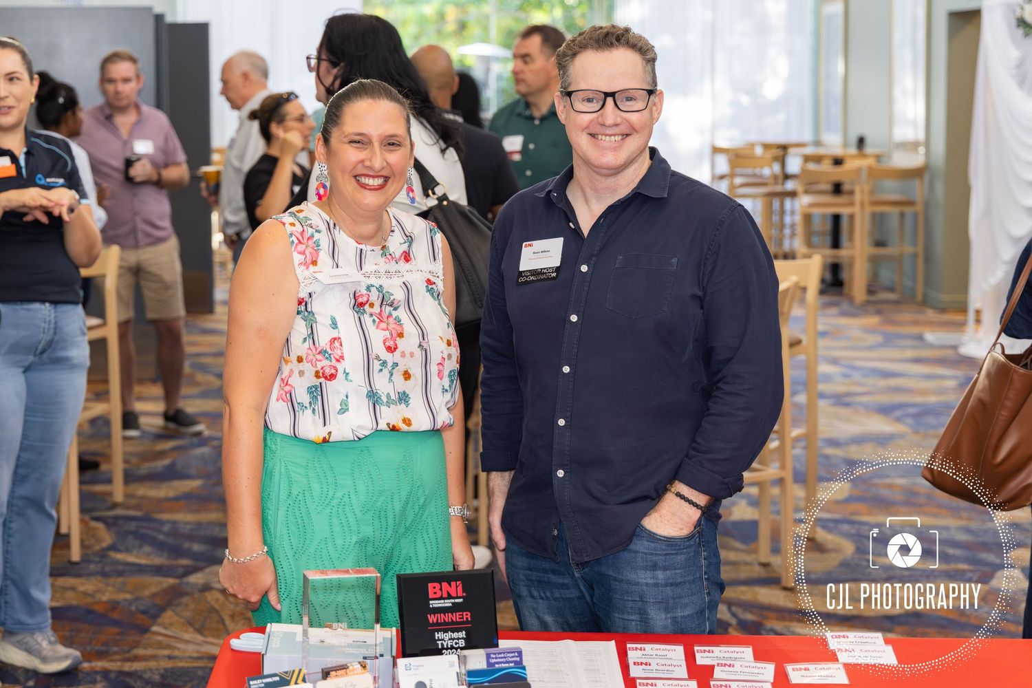 Two smiling professionals pose together at an indoor networking event beside a display table with promotional materials.