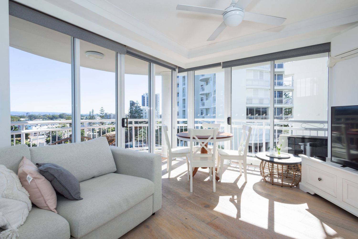 Bright modern living room with floor-to-ceiling windows, city views, ceiling fan, sofa, and dining area on a sunny day.
