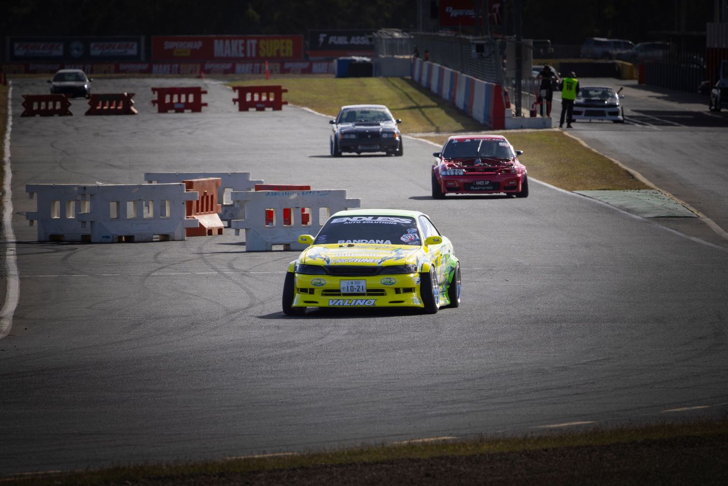Yellow race car leads a pack of cars on a circuit track during a motorsport racing event.