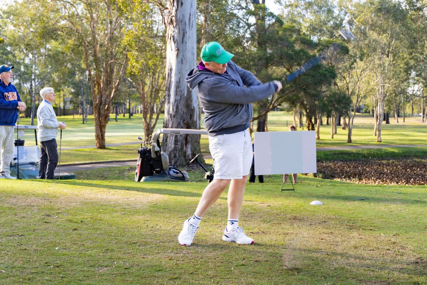 Golfer in green cap completing a full swing on a tree-lined fairway during a sunny outdoor golf tournament.
