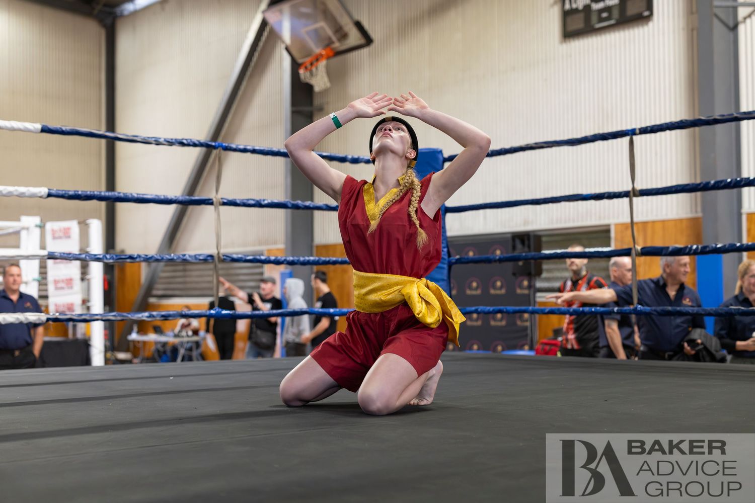Boxer kneeling on ring canvas in red and yellow shorts, hands raised, during a boxing match at an indoor venue.