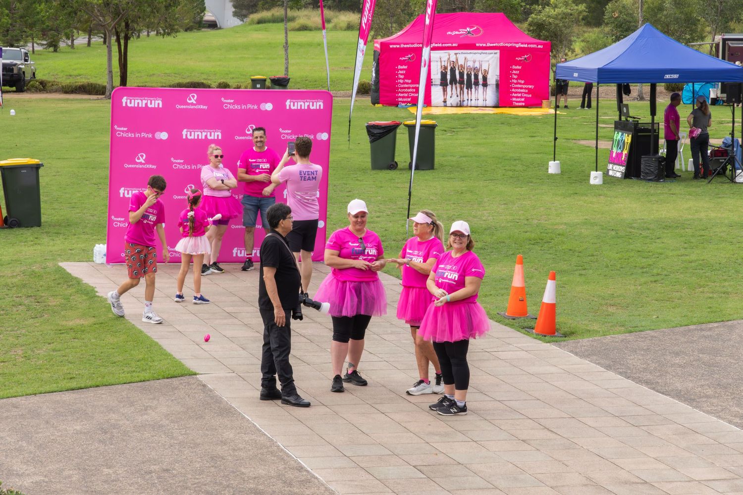 Women in pink outfits gather at a fun run event with pink signage, tents, and orange cones on a green lawn.