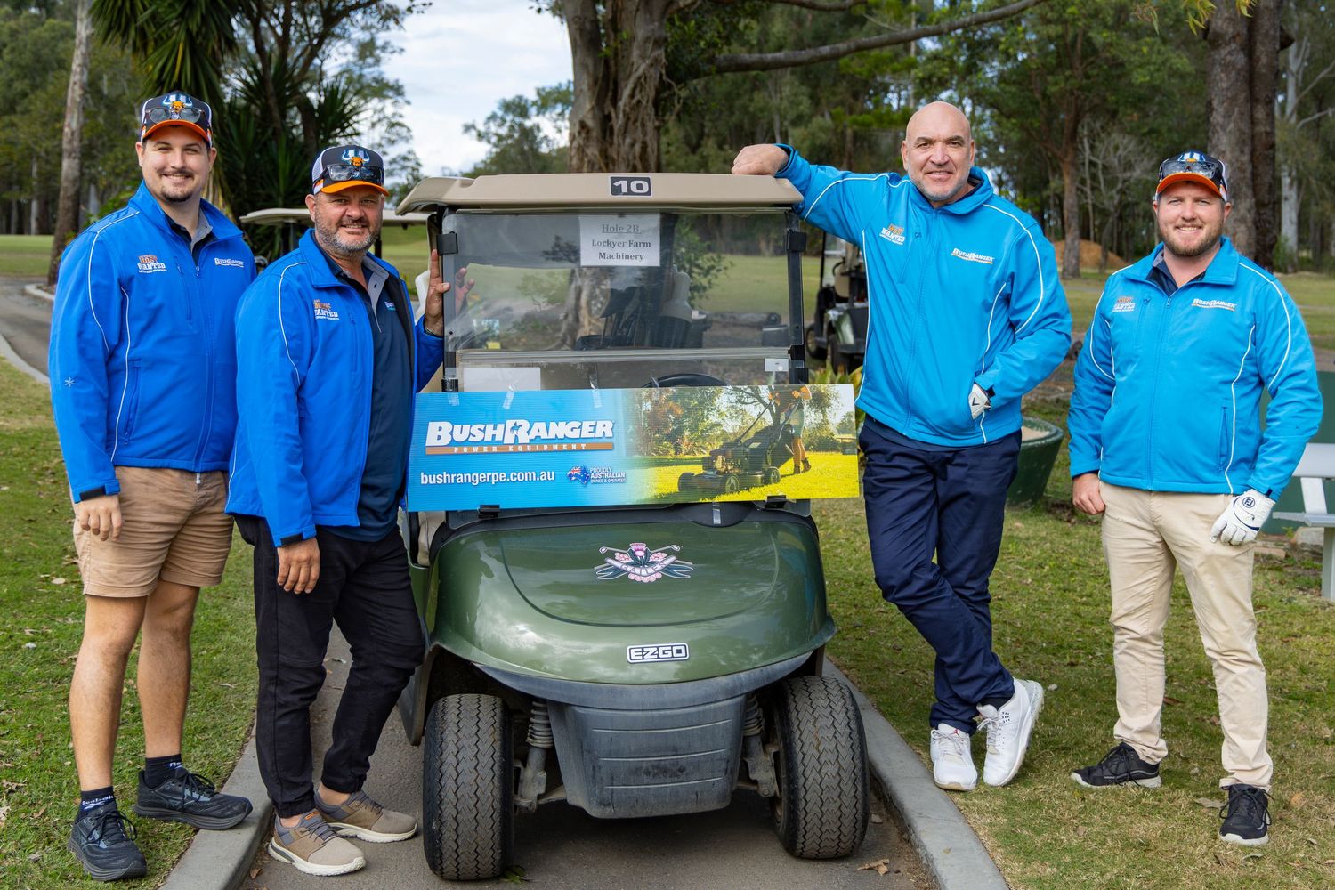 Four men in blue jackets posing beside a BushRanger-branded green golf cart on a tree-lined golf course path.