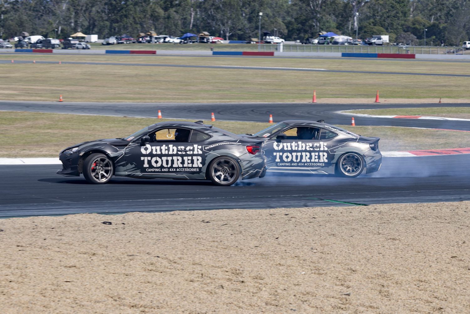Two Outback Tourer drift cars competing on a racing circuit, leaving tire smoke trails on the track.
