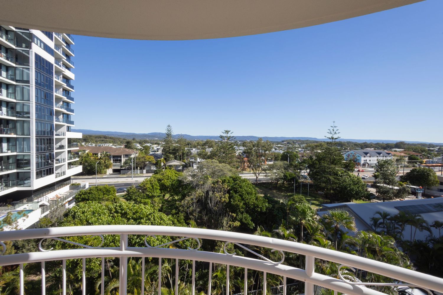 Curved white balcony railing with panoramic views of lush treetops, coastal suburb, and mountain ranges under a clear blue sky.
