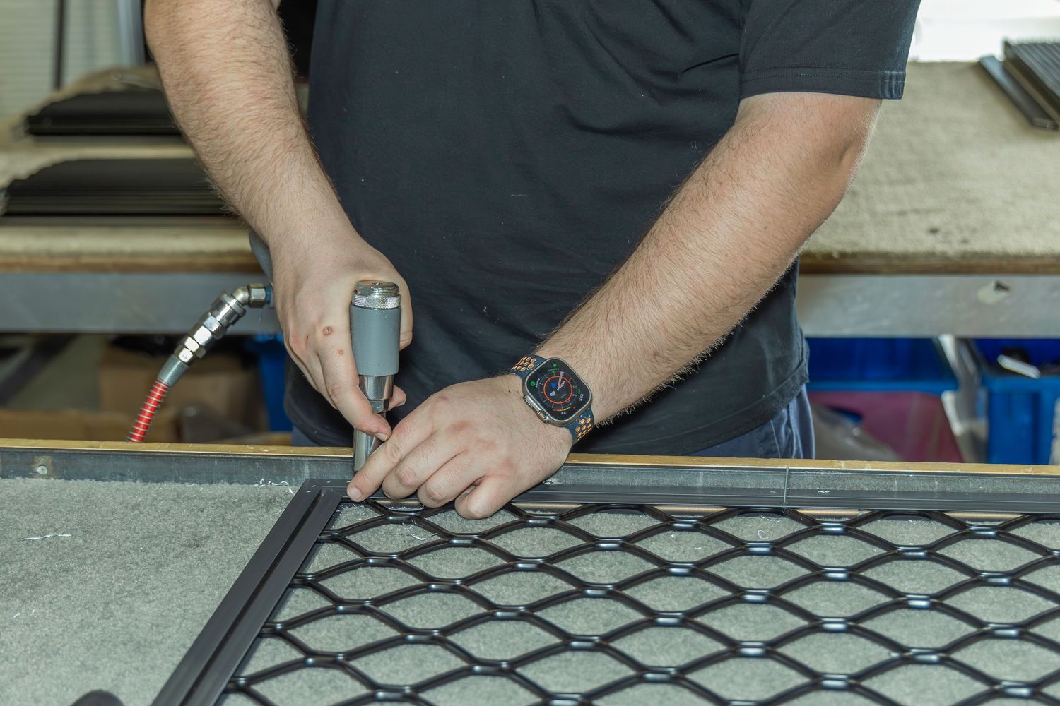 Close-up of a man using a power screwdriver to assemble a metal mesh panel in a workshop.