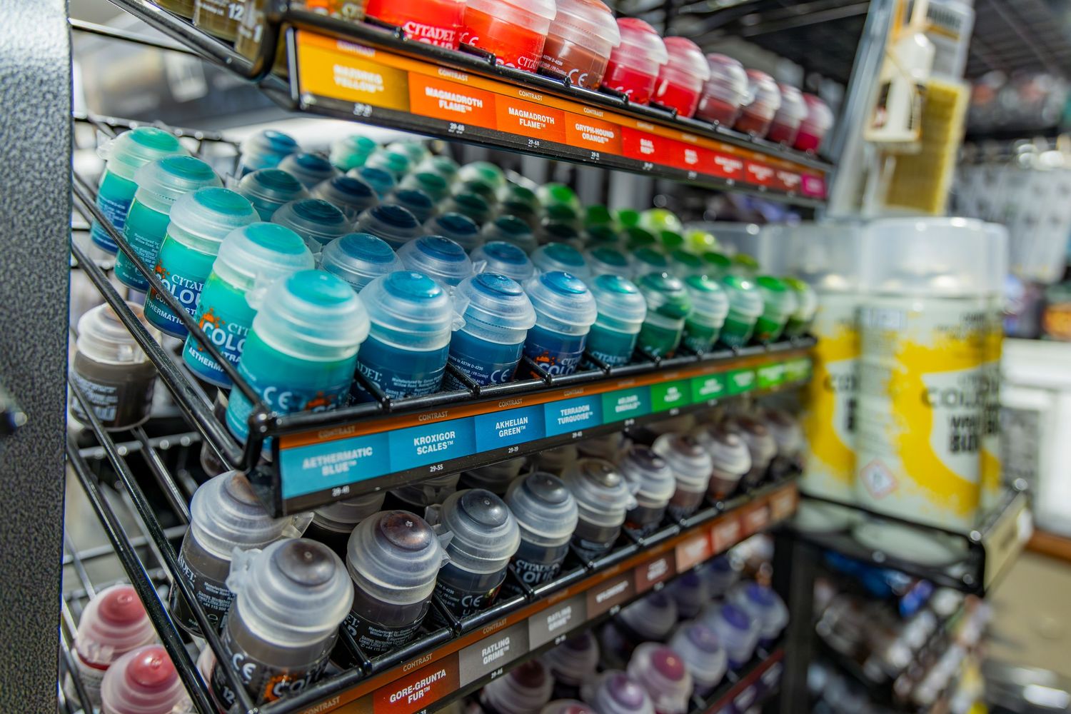 Rows of colorful Citadel hobby paint pots displayed on black wire shelving racks in a game store.