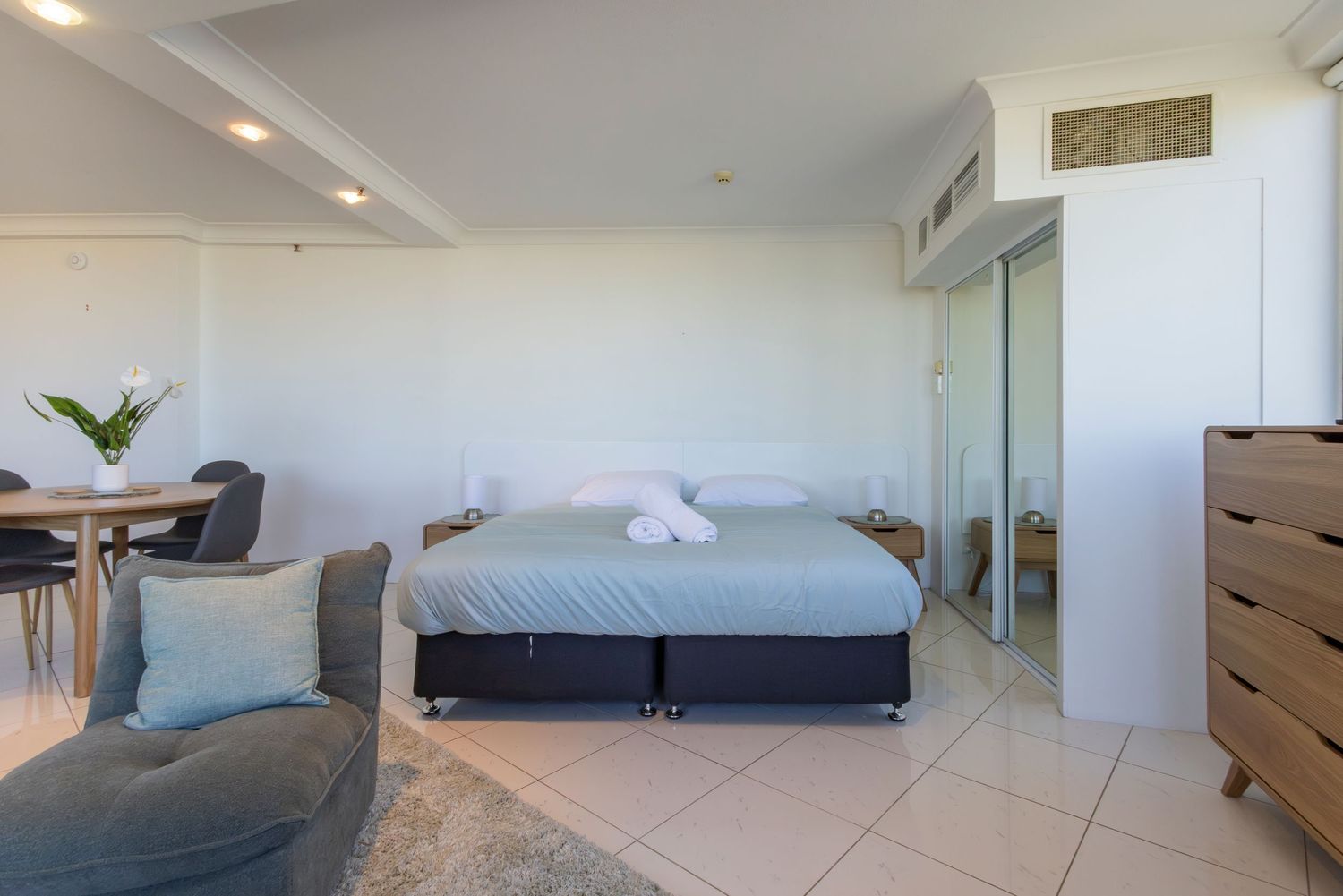 Modern studio apartment with queen bed, grey armchair, dining area, and white walls bathed in natural light.