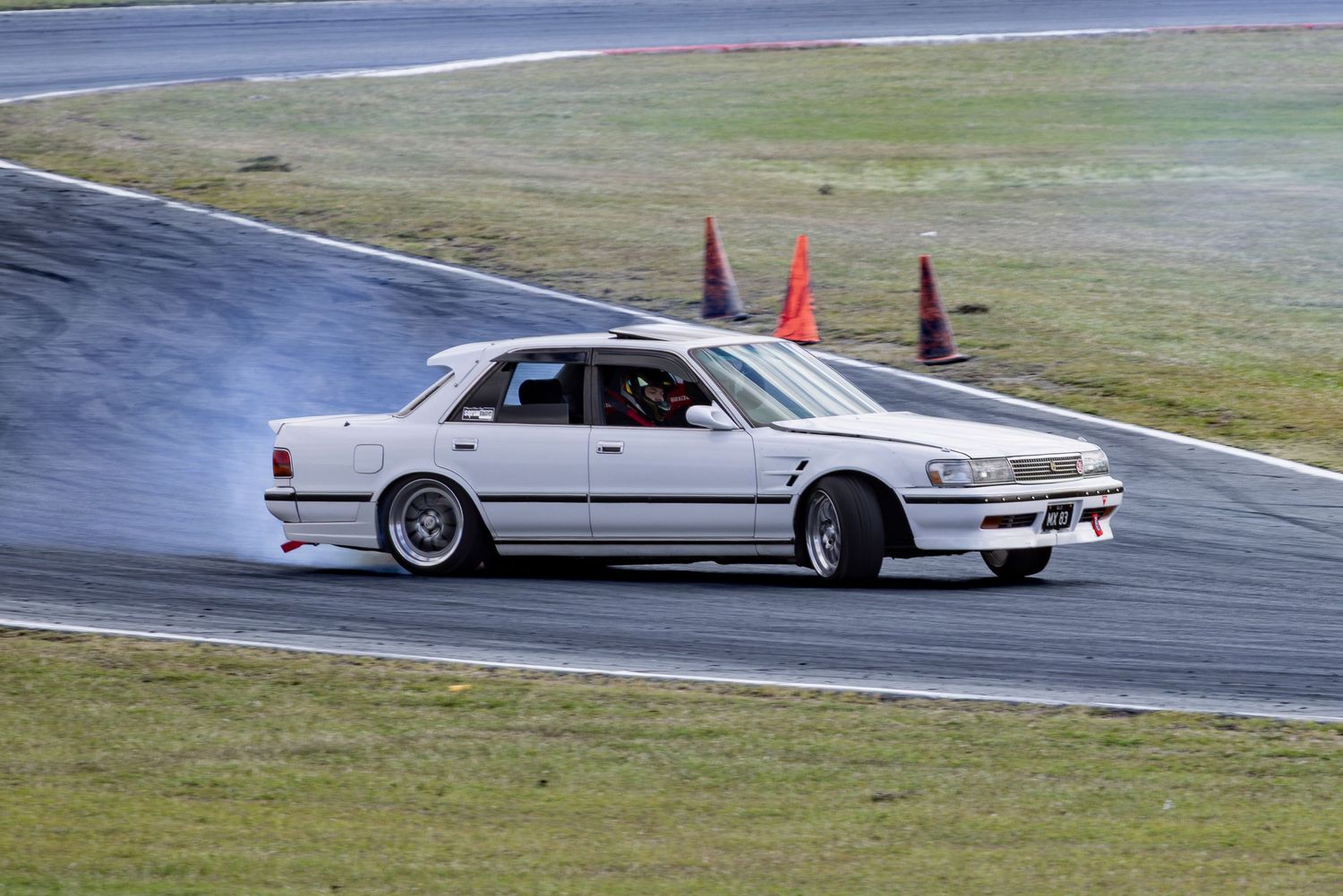 White sedan drifting on a race track with smoke from tires, orange cones visible in the background.