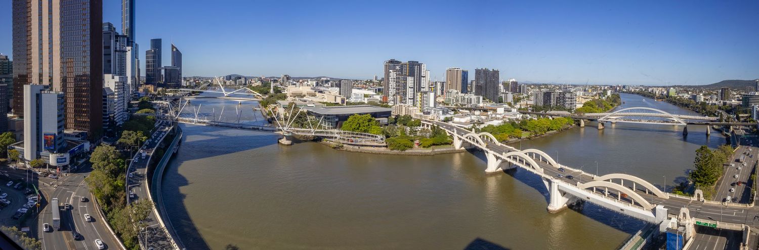 Aerial panoramic view of Brisbane River winding through the city CBD with bridges, parklands, and skyline on a clear day.