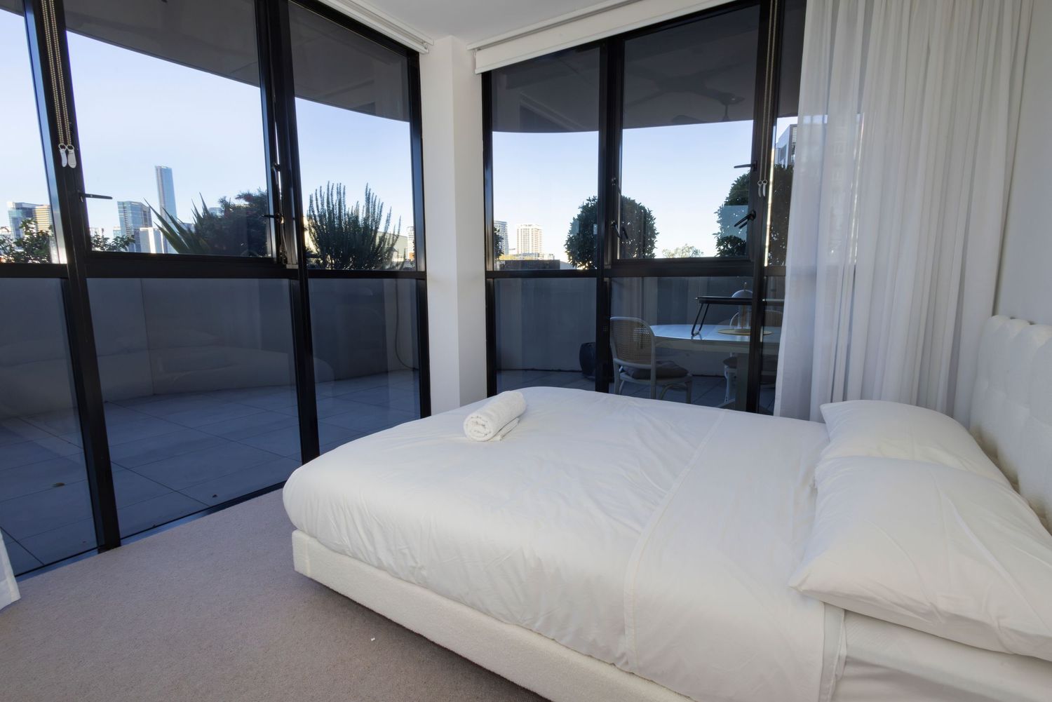 Modern white bedroom with floor-to-ceiling windows offering city views and access to a private balcony.