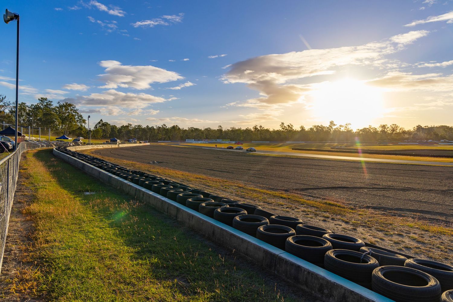 Sunset over a dirt racing track lined with tires, surrounded by trees under a blue sky with scattered clouds.